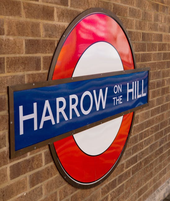 A close-up view of a classic London Underground sign mounted on the exterior brick wall of a building, featuring the iconic red circle with a blue bar and white lettering. The sky above is partly clou
