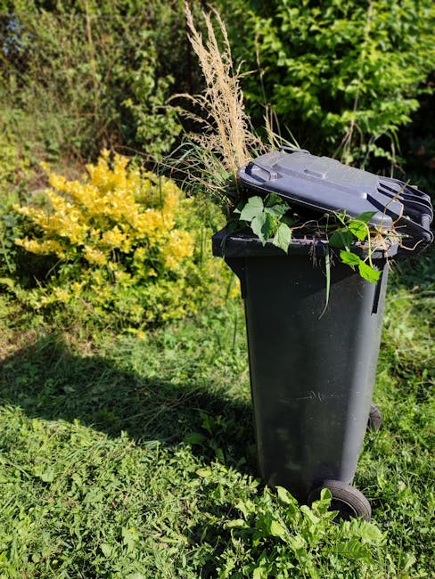 A green outdoor waste bin with a rounded lid filled with assorted garden waste and litter, wrapped in a blue plastic bag, situated on a grassy area within a landscaped garden. The background features 